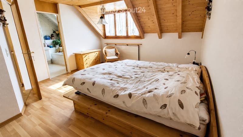 Cozy attic bedroom with wooden floors, slanted wooden ceiling, large window, bed with leaf-patterned duvet, wicker chair, nightstand, and lamp.