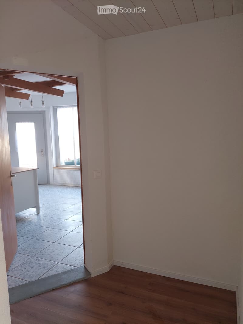 Empty hallway with wooden flooring and light pink walls, leading to an adjacent room with tiled flooring and white walls.