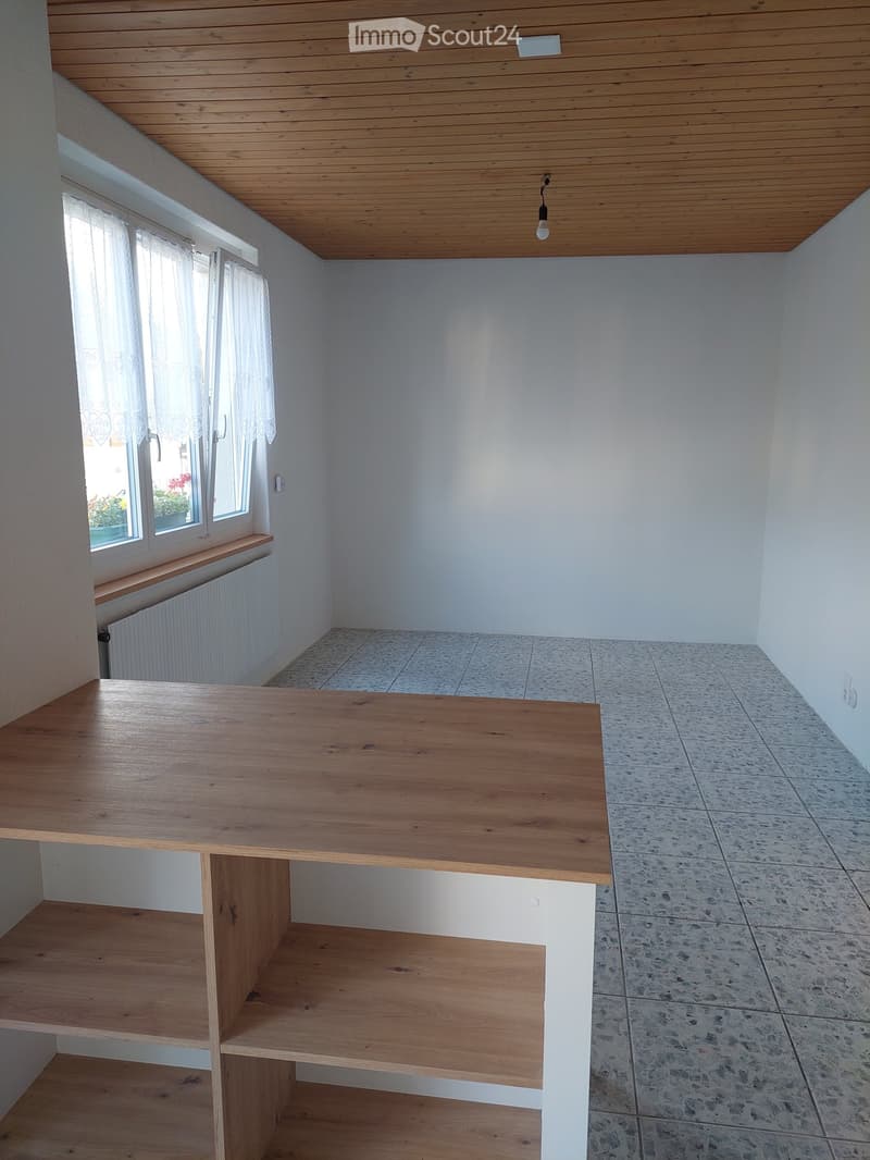 A small, unfurnished room with a wooden table and shelves, white walls, and a window with curtains. The floor is tiled, and the ceiling is wooden.