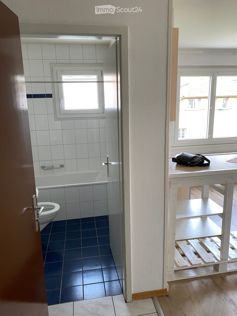 A small, partially furnished room with a white table holding a black backpack, a wooden shelf, and an open doorway leading to a tiled bathroom with a toilet and bathtub.