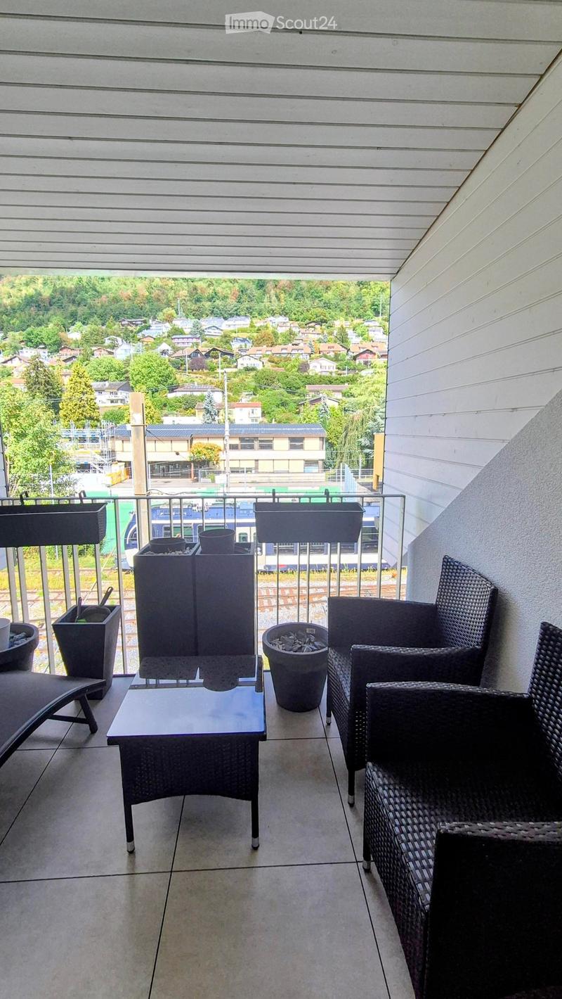 Balcony with a view of a town, featuring a glass table, wicker chairs, and several decorative pots.