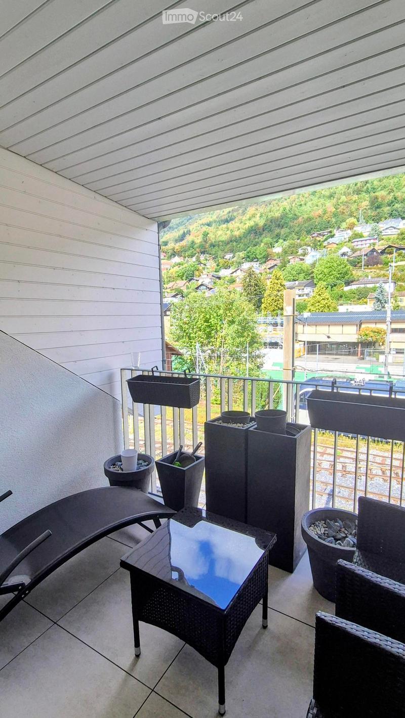 Balcony with a view of a town and surrounding greenery, featuring a glass-topped table, wicker chairs, a sun lounger, and several decorative pots.