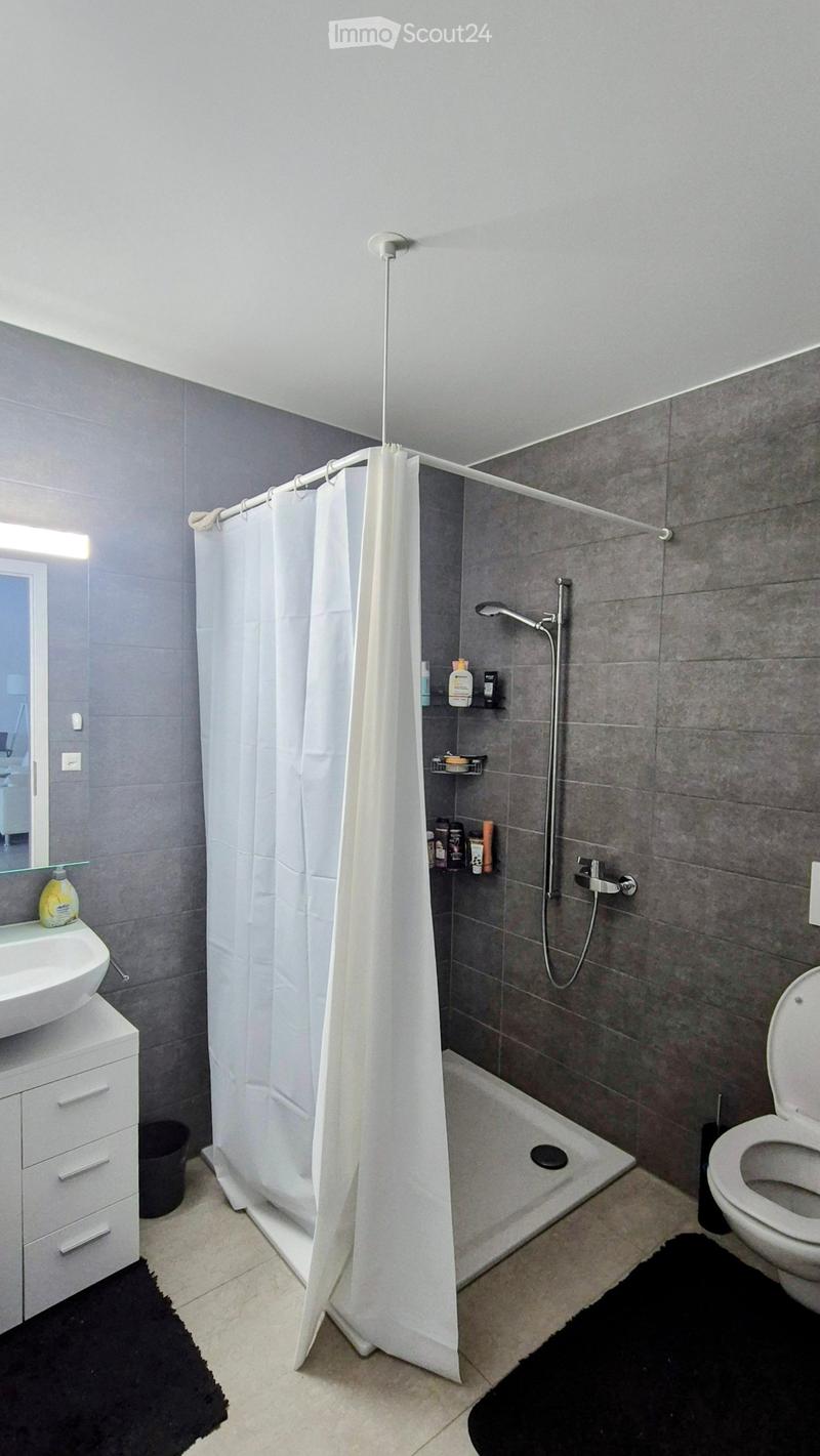 Modern bathroom with grey tiled walls, white shower curtain, sink with mirror, toilet, and shower area with a handheld showerhead.