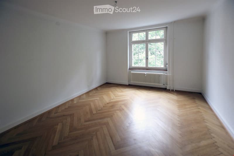 Empty room with white walls, wooden herringbone flooring, radiator, and large window with trees visible outside.