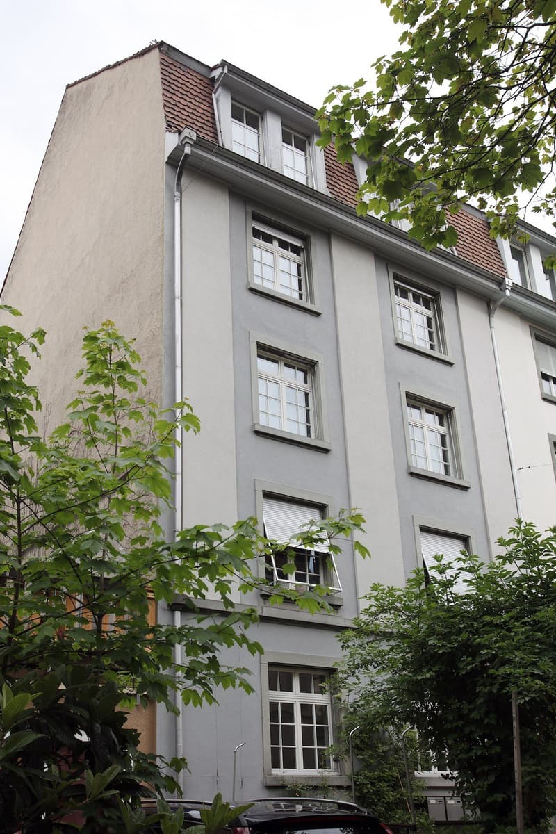 Multiple story apartment building, beige and grey painted wall, white windows, tree-lined front area