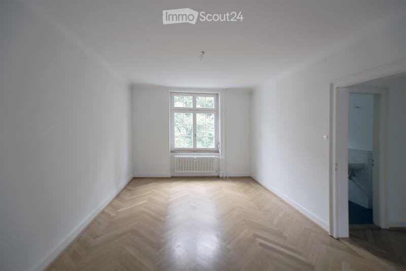 empty room, wooden floors, white walls, large window with white frames, radiator, open door leading to bathroom