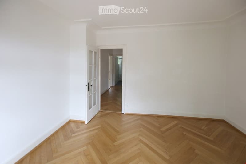 Empty room with white walls and herringbone wooden flooring, featuring an open doorway leading to another room.