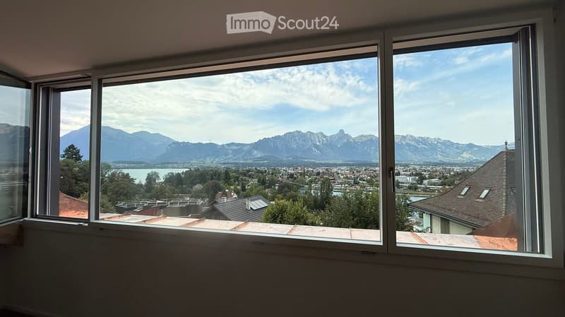 Empty room with large windows offering a scenic view of mountains and a town below.