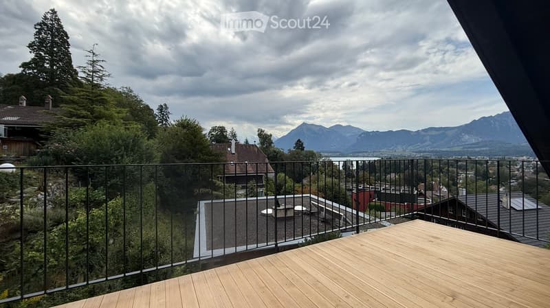 Wooden balcony with metal railing, overlooking a scenic landscape of trees, houses, and distant mountains.