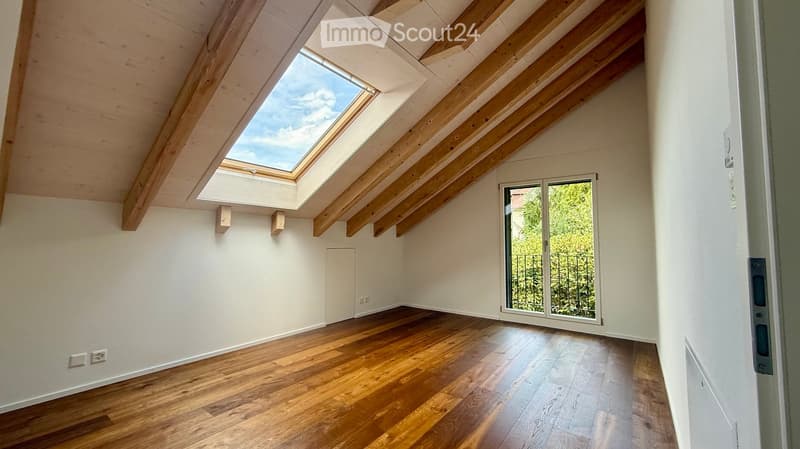 Bright, empty attic room with wooden flooring, white walls, skylight, and large window with a view of greenery.