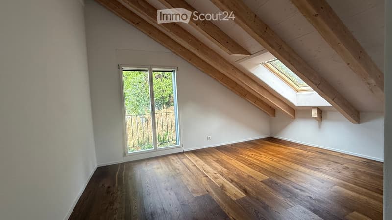 Empty attic room with wooden beams, wooden flooring, large skylight, and a window with a view of greenery.