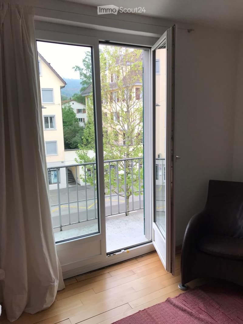 A modern room with wooden flooring, a large sliding glass door opening to a balcony with a view of the city and mountains, a single chair, and a red rug.