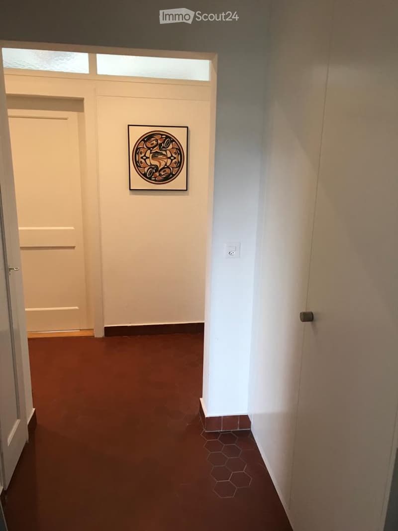 A narrow hallway with a brown carpeted floor, white walls, and a framed circular artwork on one wall. The hallway features a door at one end and a small window above it, with a light switch visible on the right wall.