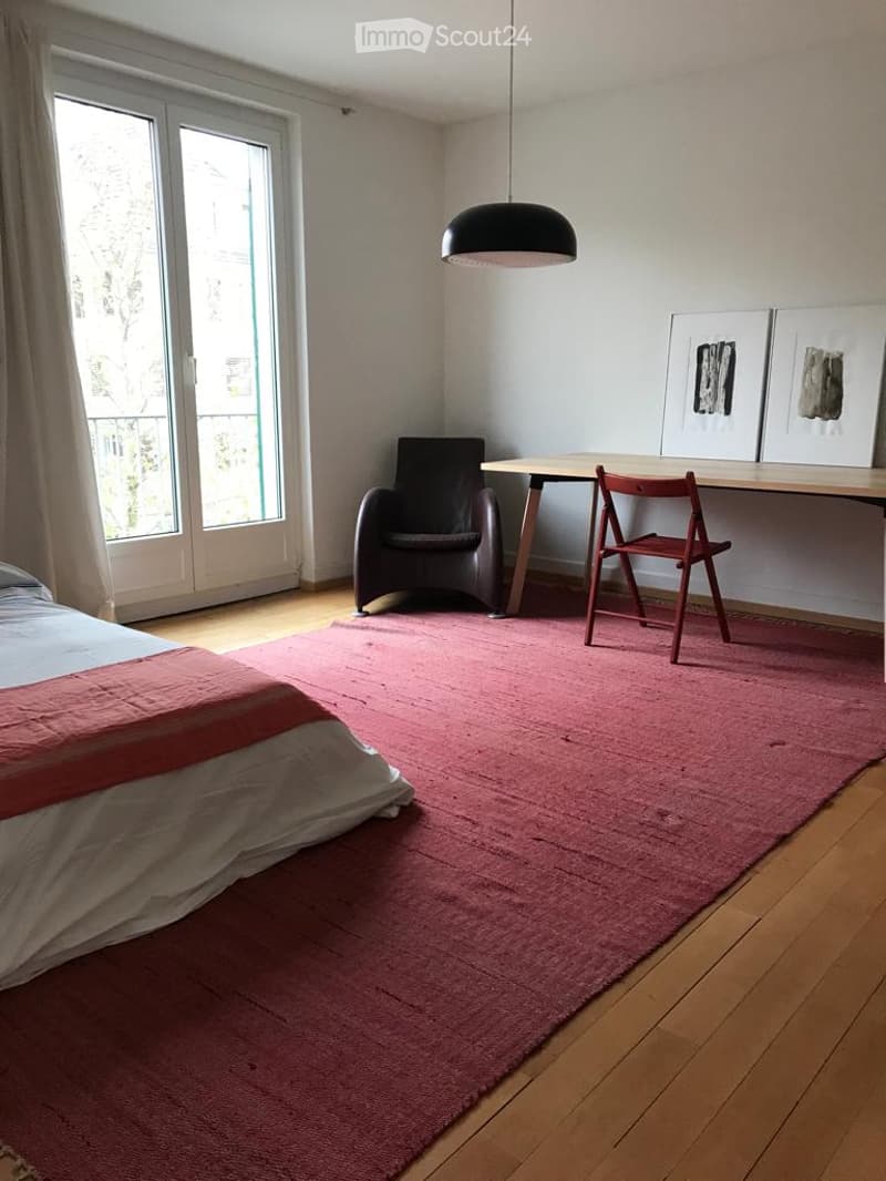 A modern bedroom with a wooden floor, a large red rug, a bed with white and red bedding, a black chair, a wooden table, a red chair, a hanging lamp, and two picture frames on the wall.