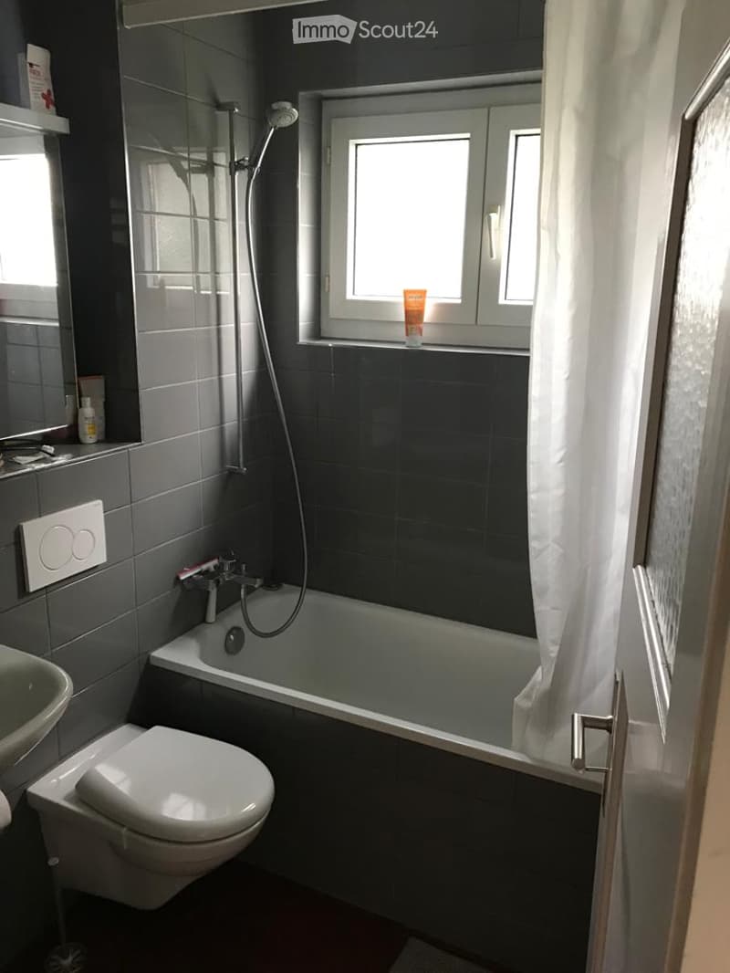 Modern bathroom with dark grey tiles, a bathtub, toilet, sink, shower, and a window with a white curtain.