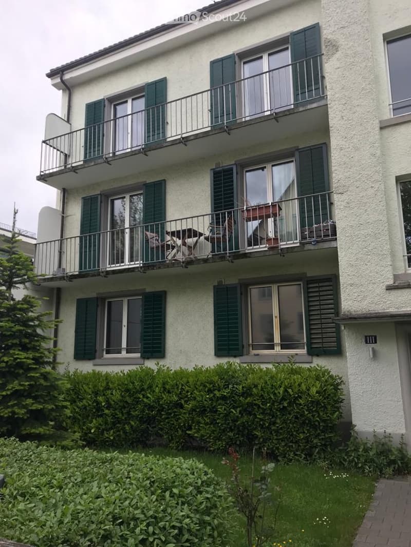 Three-story residential building with white facade, green shutters, and multiple balconies. Balconies feature outdoor furniture and potted plants.