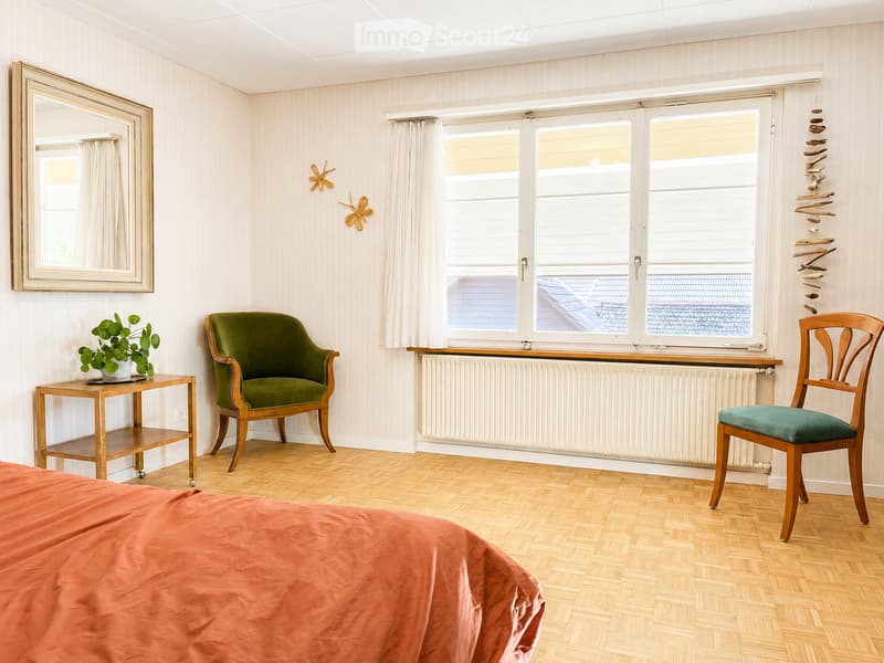 Bright bedroom with wooden flooring, large window with white curtains, green armchair, small wooden table with plant, and a mirror on the wall.