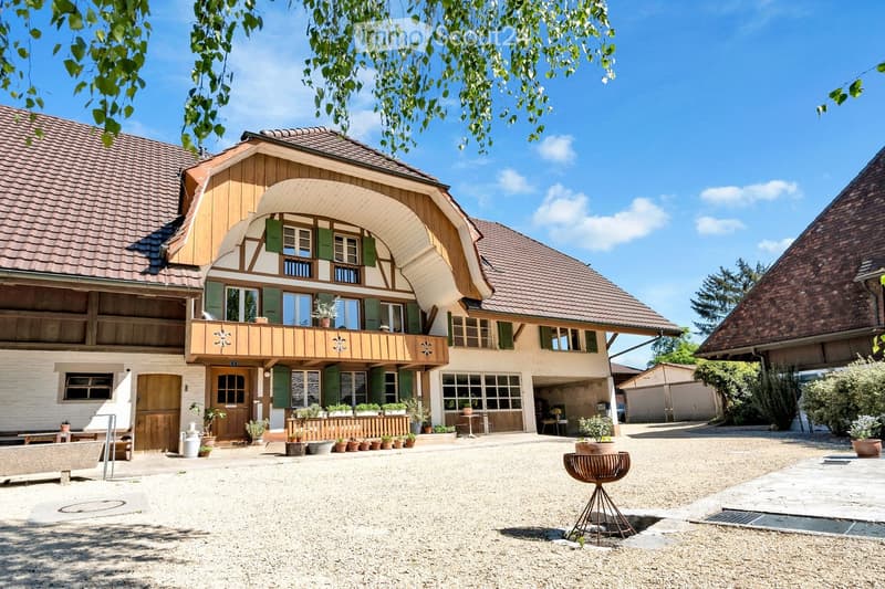 A two-story traditional house with a wooden facade, green shutters, and a covered balcony. The house features multiple windows, a wooden door, and a gravel driveway with potted plants and outdoor furniture.