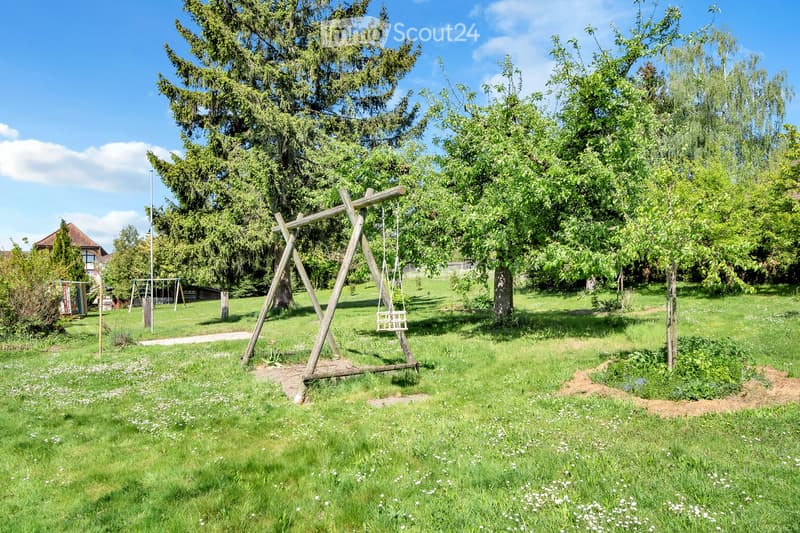 A well-maintained garden with a wooden swing set, lush green grass, and several tall trees providing shade. The area is dotted with small white flowers and features a small wooden playhouse in the background.