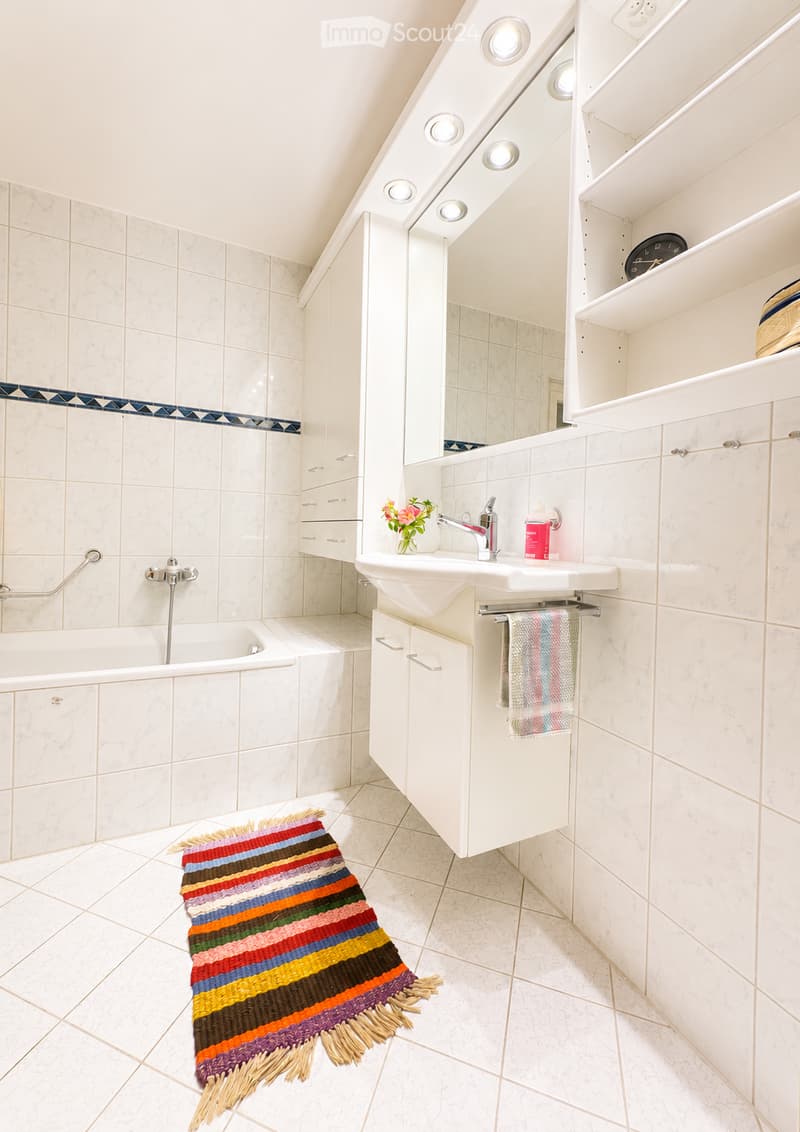 Modern bathroom with white tiles, a bathtub, a sink with a mirror, a colorful striped rug, and built-in cabinets.