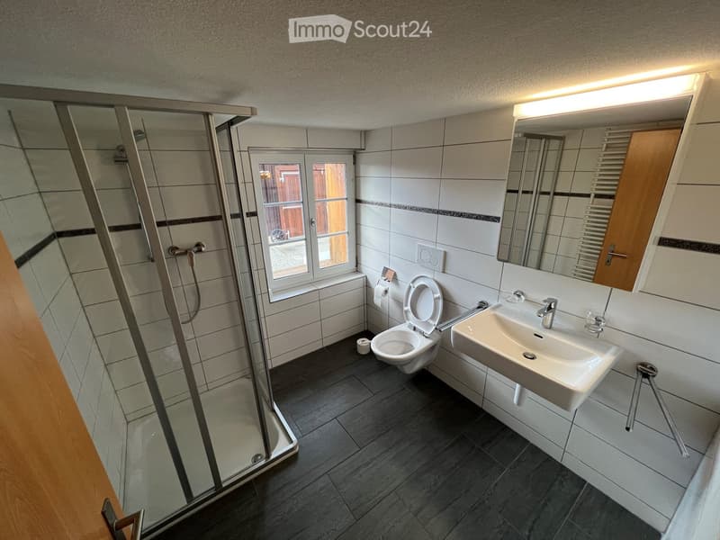 white tiled walls, black tiled floor, glass shower enclosure, white sink, toilet, mirror, window