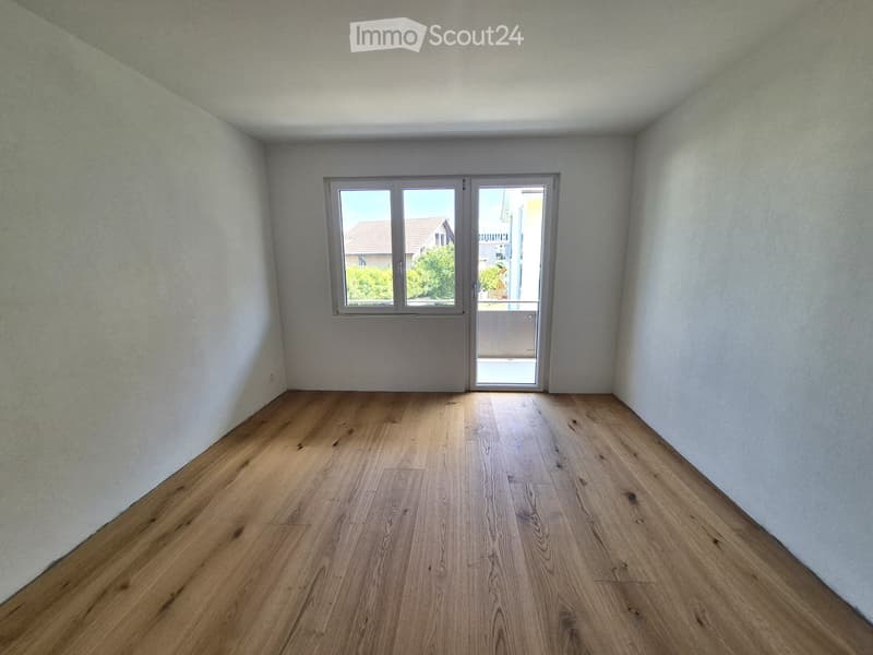 Empty room with light wooden flooring and white walls, featuring a large window and a sliding glass door leading to a balcony.