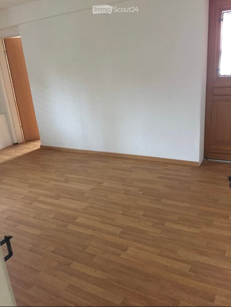 Empty room with wooden flooring and white walls, featuring a wooden door and a small window.