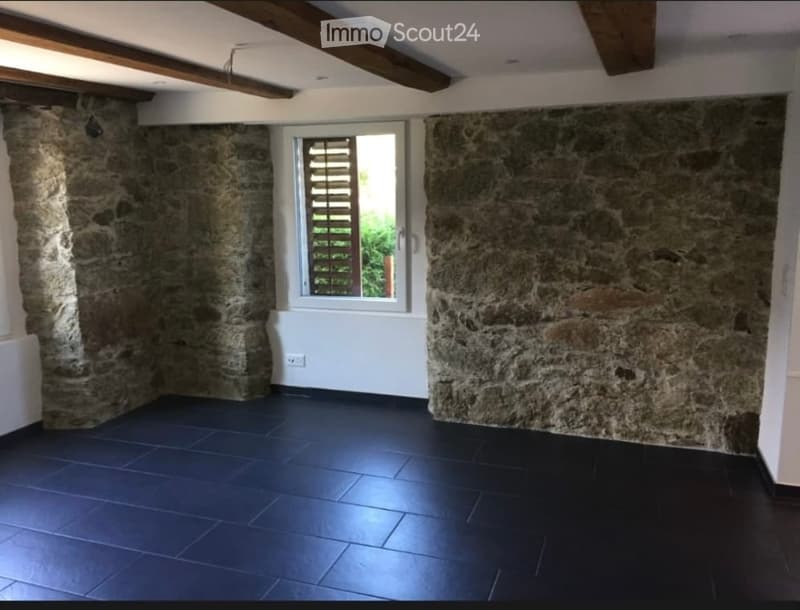 Empty room with exposed stone walls, dark tiled floor, and a single window with shutters.