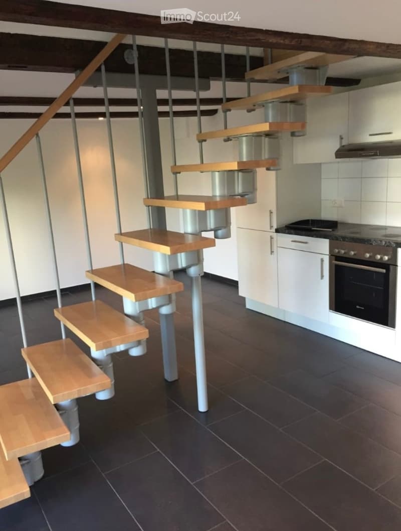 Modern kitchen with dark tiled floor, white cabinets, built-in oven, and a unique staircase with wooden steps and metal supports.