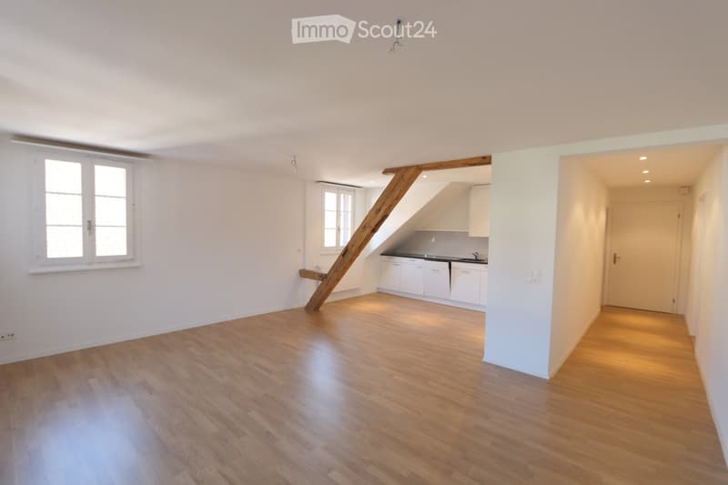 Empty room with wooden flooring, white walls, and large windows. A wooden staircase leads to an adjacent kitchen area with white cabinets and a sink.