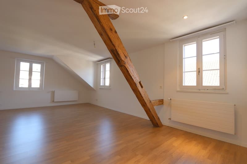 Empty attic room with wooden flooring, white walls, multiple windows, and exposed wooden beam. No furniture or equipment present.