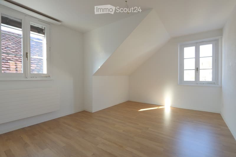 Empty attic room with wooden flooring, white walls, and two windows offering views of the roof.