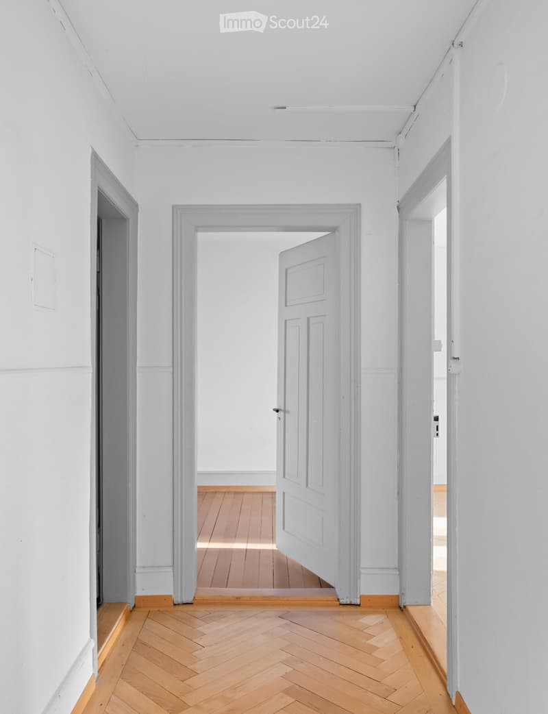 Empty hallway with white walls, wooden herringbone flooring, and three open doors leading to other rooms.