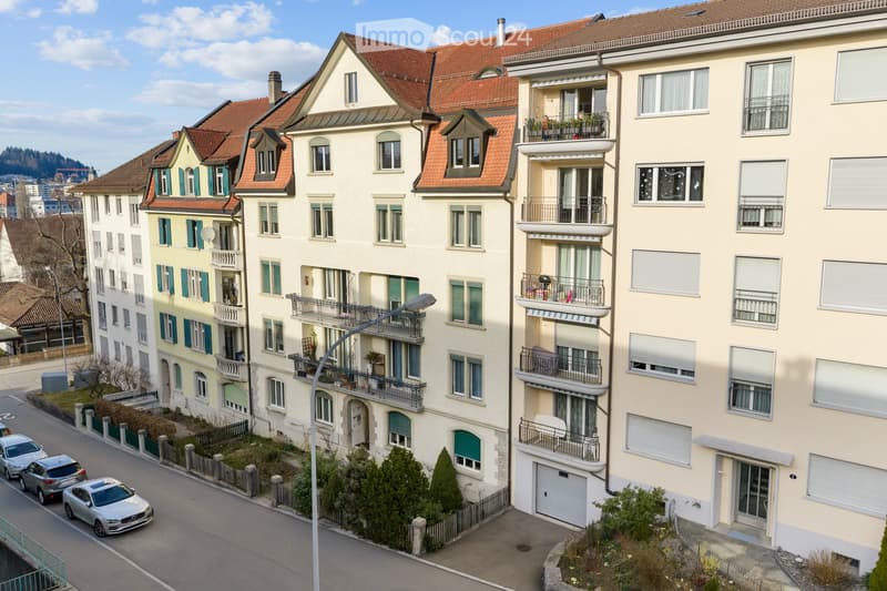 A multi-story residential building with balconies, large windows, and a red-tiled roof. The building is surrounded by other similar structures, with parked cars and greenery visible in the foreground.