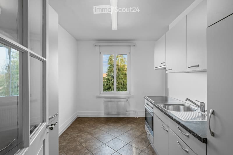Modern kitchen with white cabinets, stainless steel sink, oven, and tiled floor. Large windows provide natural light.