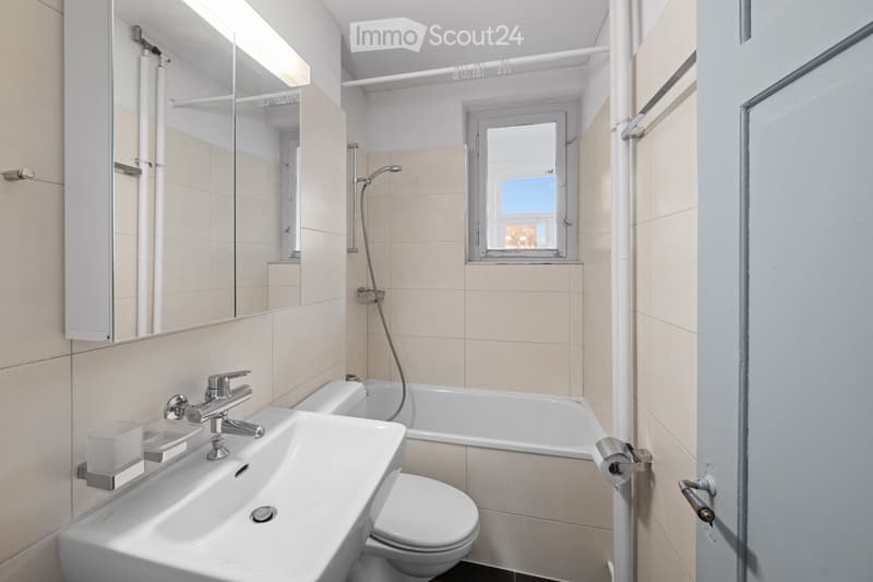 Modern bathroom with a white sink, bathtub, toilet, and a large mirror above the sink. The room features beige tiles and a small window letting in natural light.