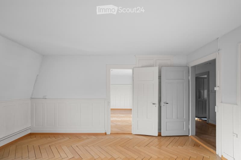 Empty room with light-colored walls, wooden herringbone flooring, and several open doors leading to adjacent spaces.