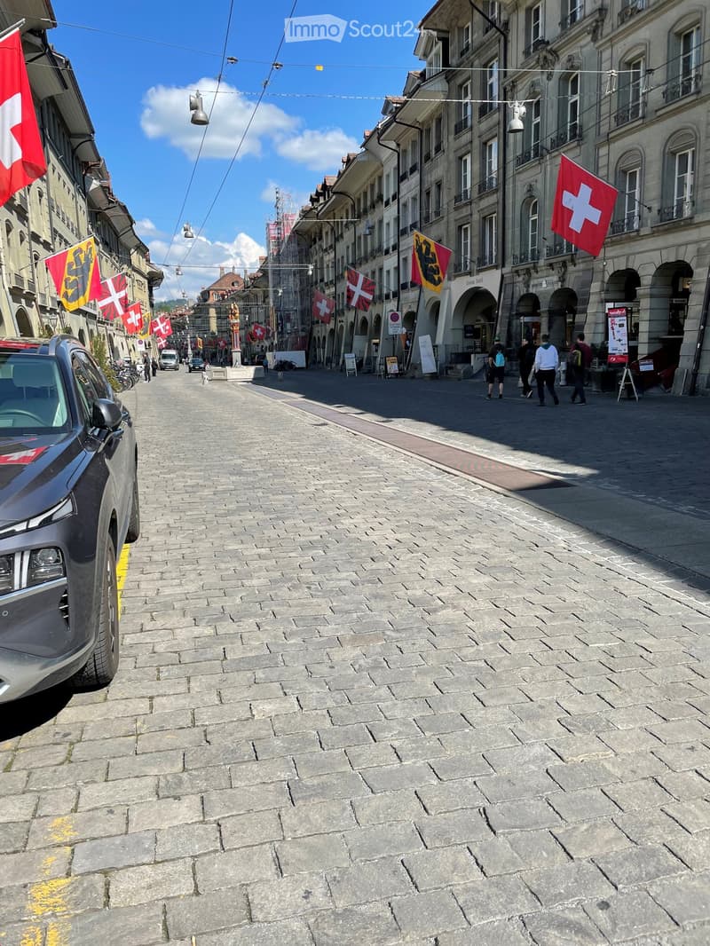 Cobblestone street with tram lines, Swiss flags, buildings, people walking, parked car