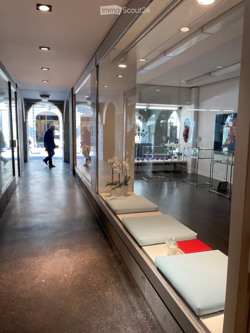 A hallway with a glass wall, a person walking, a white bench, a white flower vase, a white cushion, a red cushion, a glass table, a glass display case, a white ceiling, and a white ceiling light.