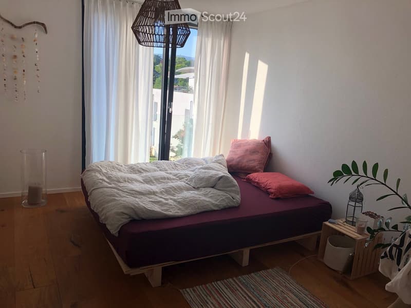 Cozy bedroom with a wooden bed frame, grey duvet, pink pillows, a small nightstand with a plant, and a hanging wicker chandelier.