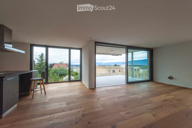 Empty room with wooden flooring, large sliding glass doors leading to a balcony, and a small kitchen area with a stool.