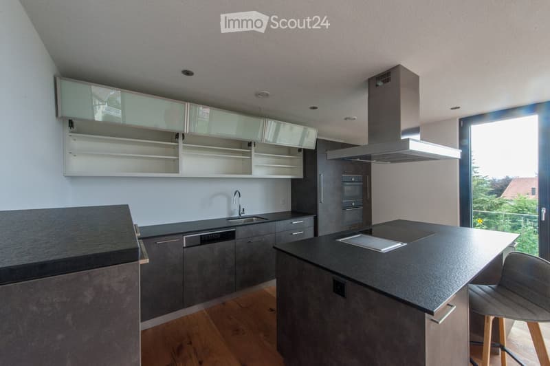 Modern kitchen with sleek black countertops, grey cabinets, built-in oven, range hood, and an island with a sink and faucet.