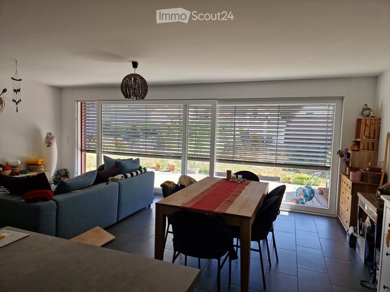 Bright living room with large windows, a sofa, a wooden dining table with chairs, and decorative items on shelves.