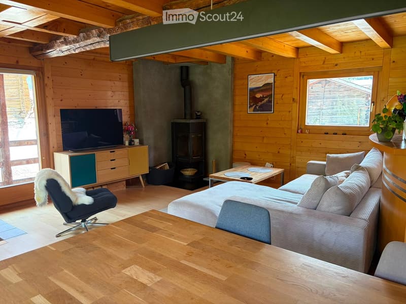 Cozy living room with wooden walls and ceiling, featuring a grey sectional sofa, coffee table, armchair with fur throw, TV on a stand, fireplace, and decorative items.