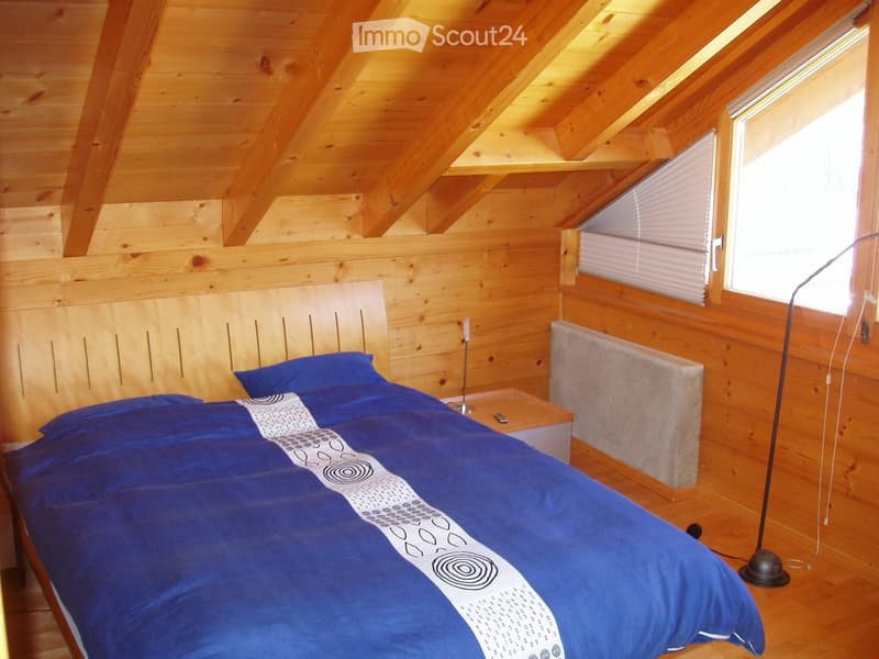 Cozy wooden bedroom with slanted ceiling, large bed with blue duvet, nightstand, floor lamp, radiator, and windows with blinds.