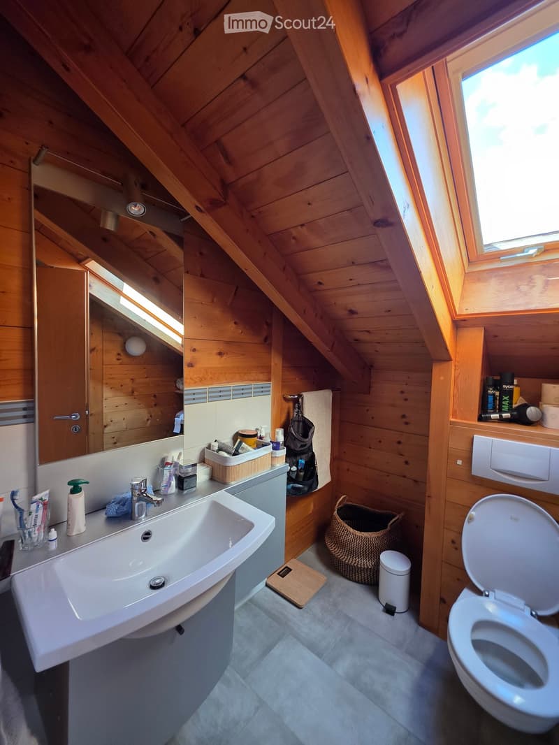 Cozy attic bathroom with wooden walls and slanted ceilings, featuring a sink, toilet, mirror, wicker basket, and various toiletries.