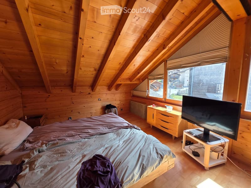 Cozy attic bedroom with wooden walls and slanted ceilings, featuring a large bed, nightstand, TV on a stand, and a small table with drawers.