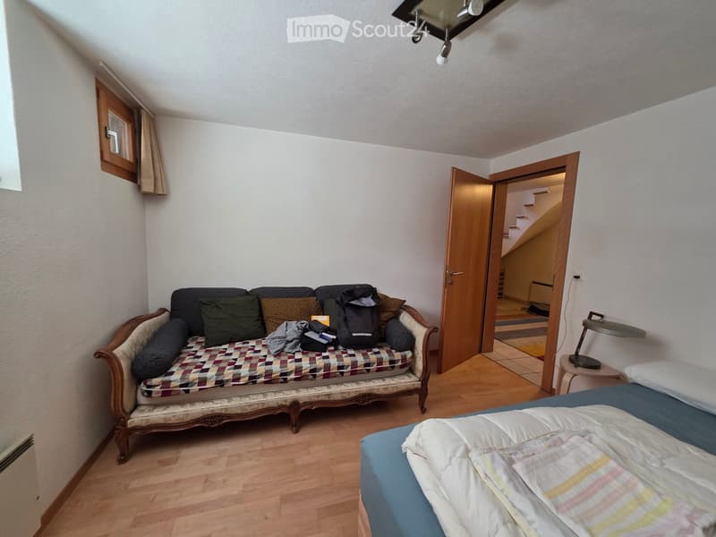 A furnished room with a sofa, bed, small table, and lamp. The room has wooden flooring and white walls.