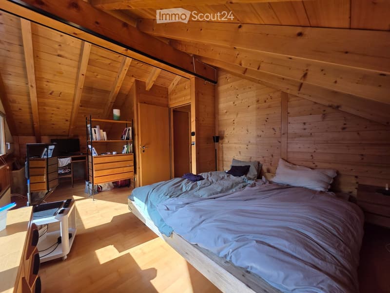 Cozy wooden bedroom with slanted ceiling, bed with blue bedding, nightstand, desk with laptop, bookshelf, and a chair.