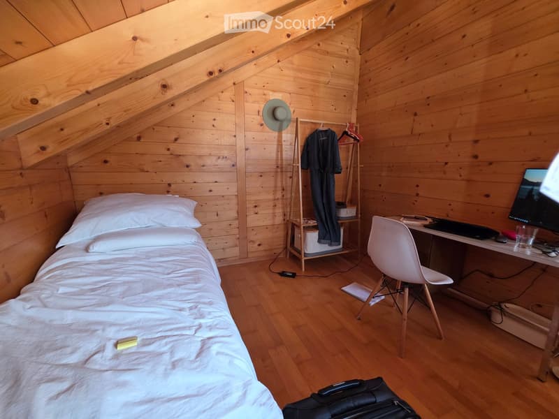 Cozy wooden bedroom with a single bed, desk, chair, and laptop. Wooden walls and slanted ceiling.